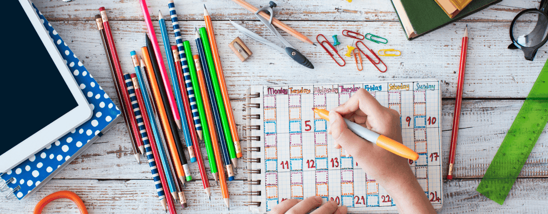 Top view of back-to-school items like pencils, a calendar, paper clips, books all on a white wood background.