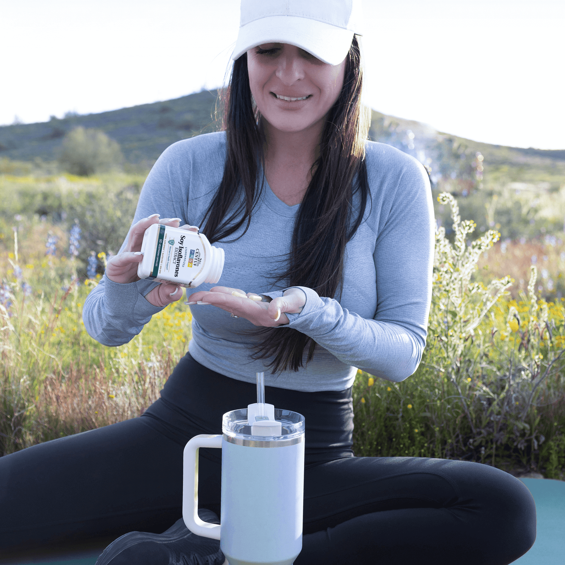 A woman on a hike in the desert sitting pouring a bottle of 21st Century Soy Isoflavones Extract vegetarian capsules with a water bottle in front.