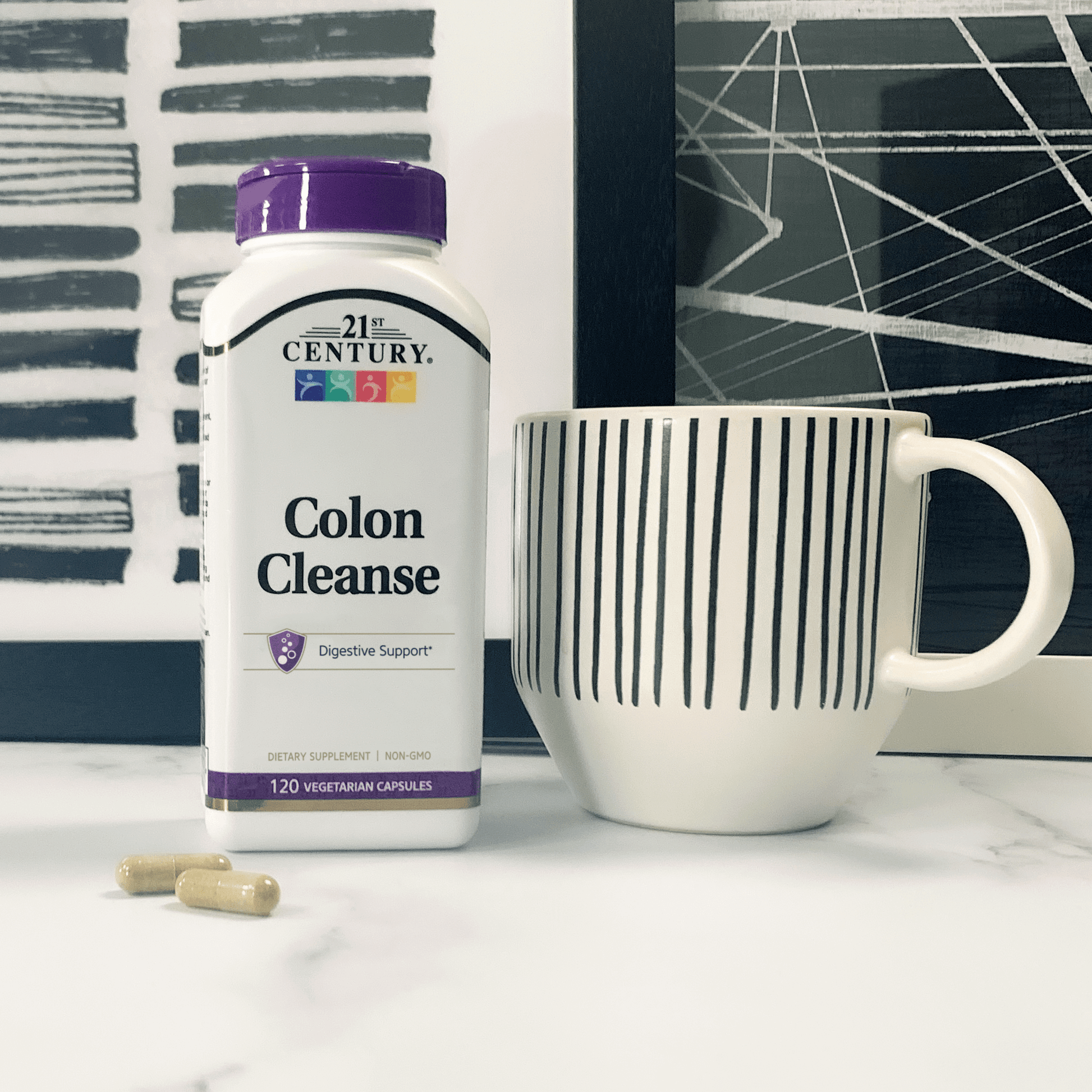 A bottle of 21st Colon Cleanse vegetarian capsules next to a coffee cup on a counter with abstract artwork in the background.