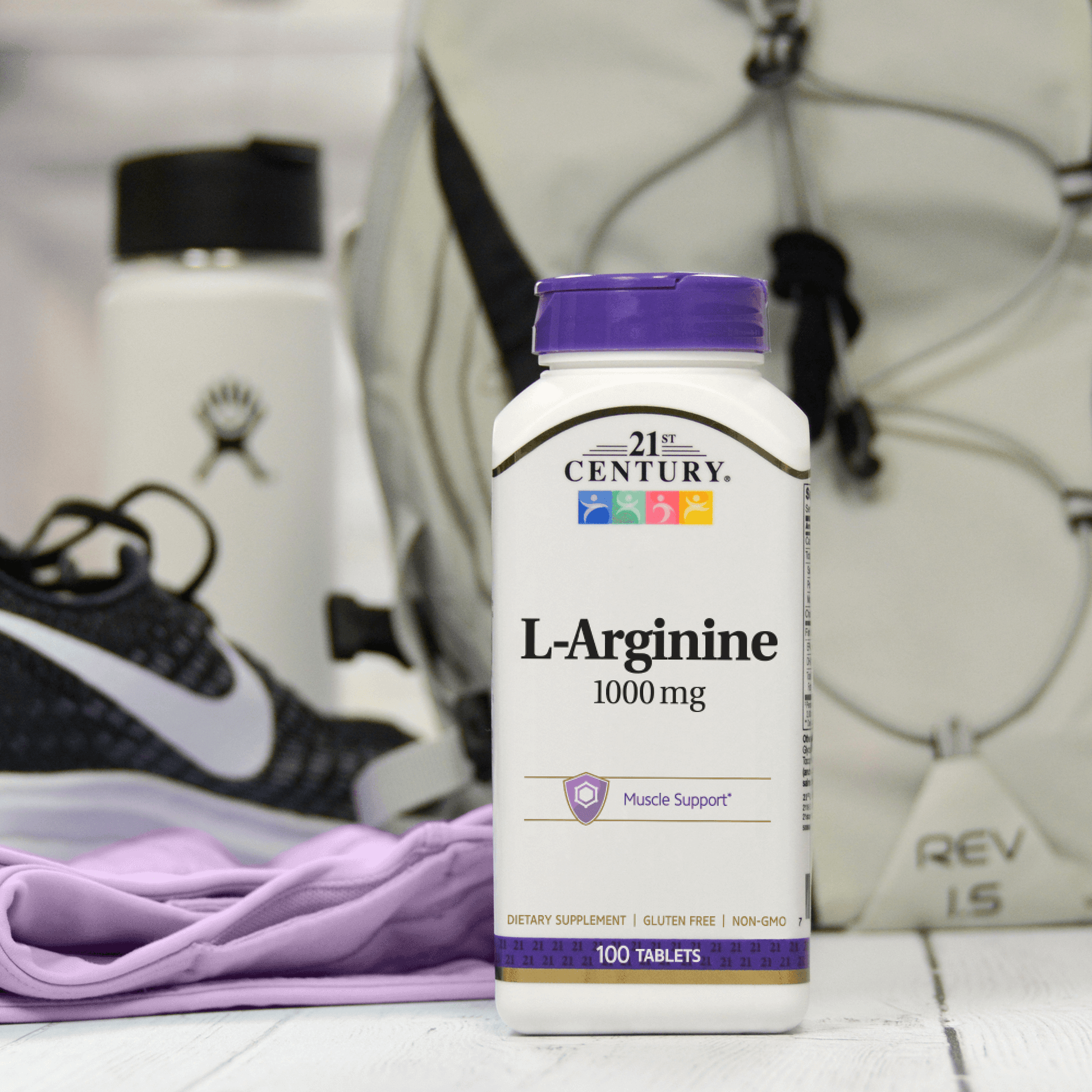 A bottle of 21st Century L-Arginine 1000 mg tablets with gym shoes, a back pack and a water bottle in the background.