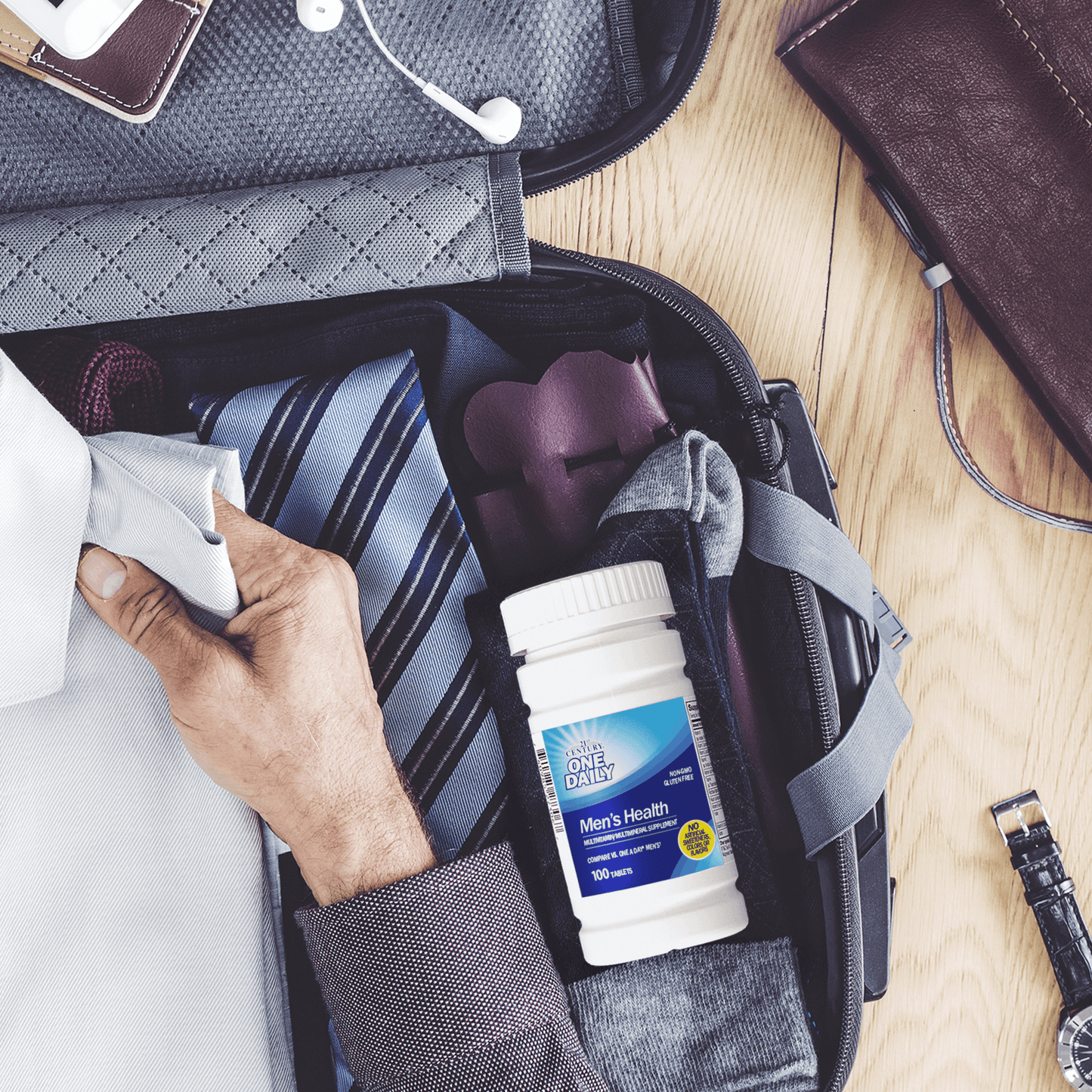 A bottle of 21st Century One Daily Men's Health Multivitamin and Multimineral tablets in a suitcase with a tie and a man placing a folded work shirt inside.