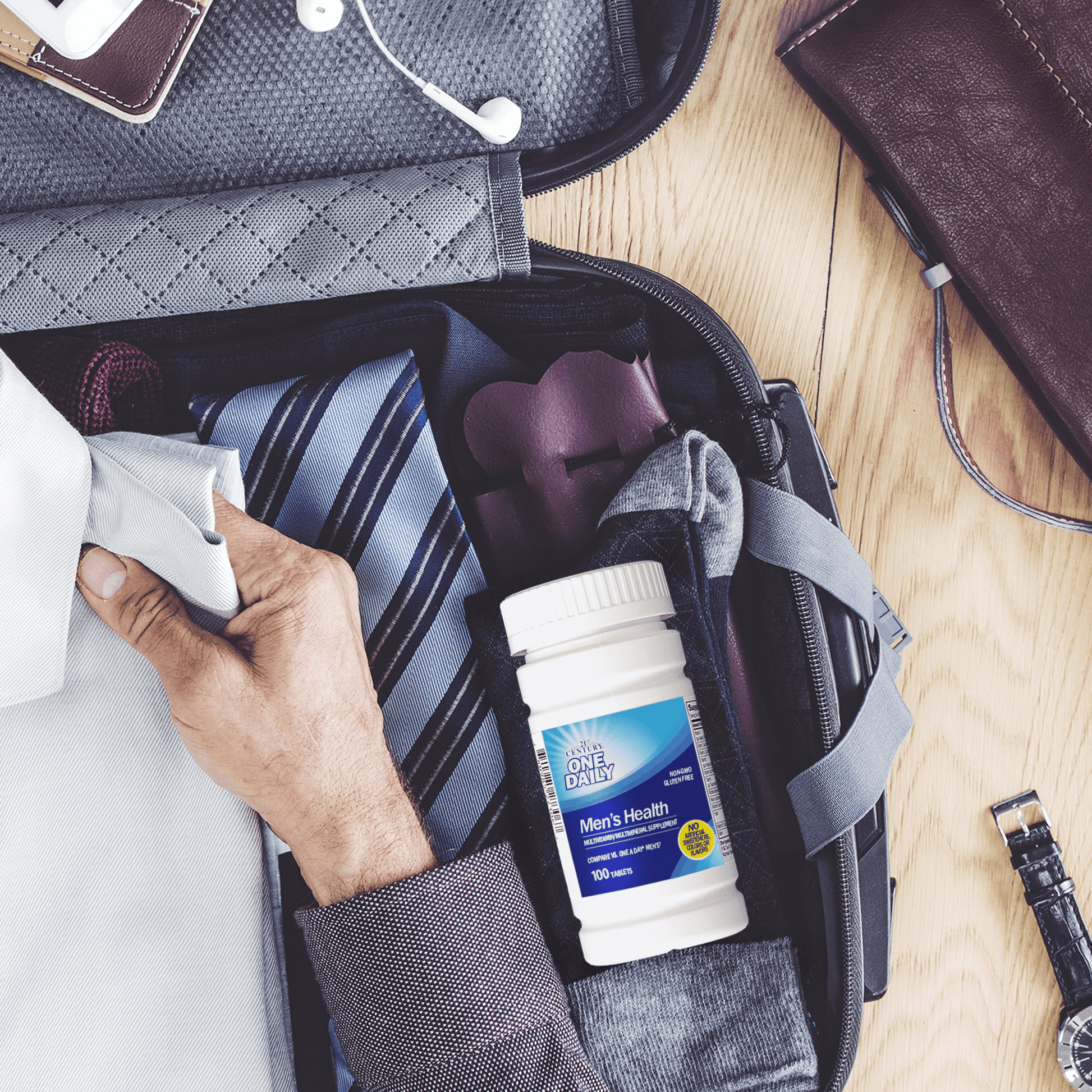 A bottle of 21st Century One Daily Men's Health Multivitamin and Multimineral tablets in a suitcase with a tie and a man placing a folded work shirt inside.