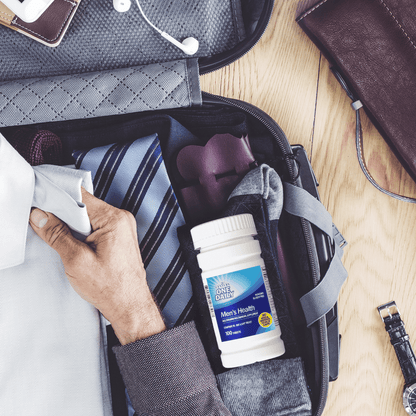 A bottle of 21st Century One Daily Men's Health Multivitamin and Multimineral tablets in a suitcase with a tie and a man placing a folded work shirt inside.