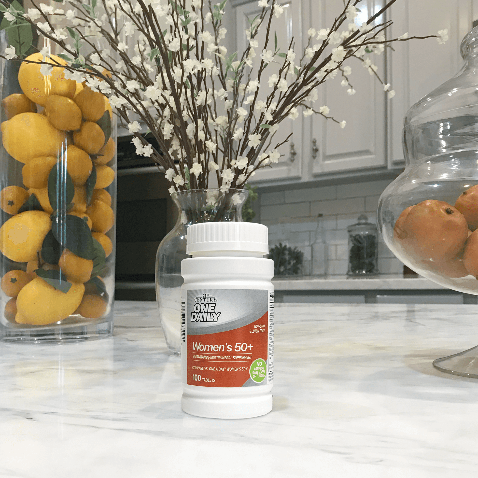 A bottle of 21st Century One Daily Women's 50+ Multivitamin and Multimineral tablets sitting on a kitchen counter with a vase of flowers and jars of fruit in the background.