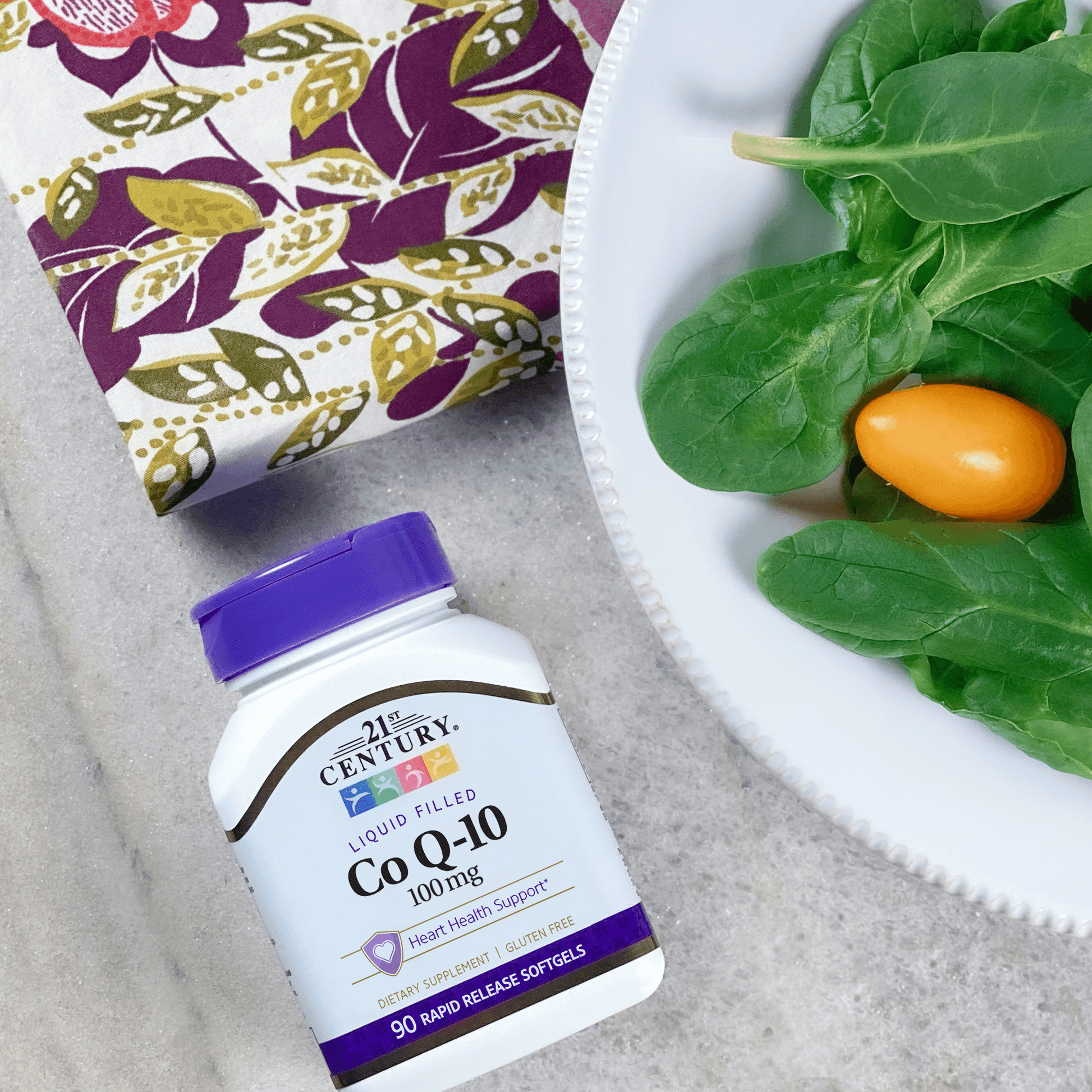 A bottle of 21st Century Co Q-10 100 mg rapid release softgels laying on a table next to a dinner plate of salad and a napkin.