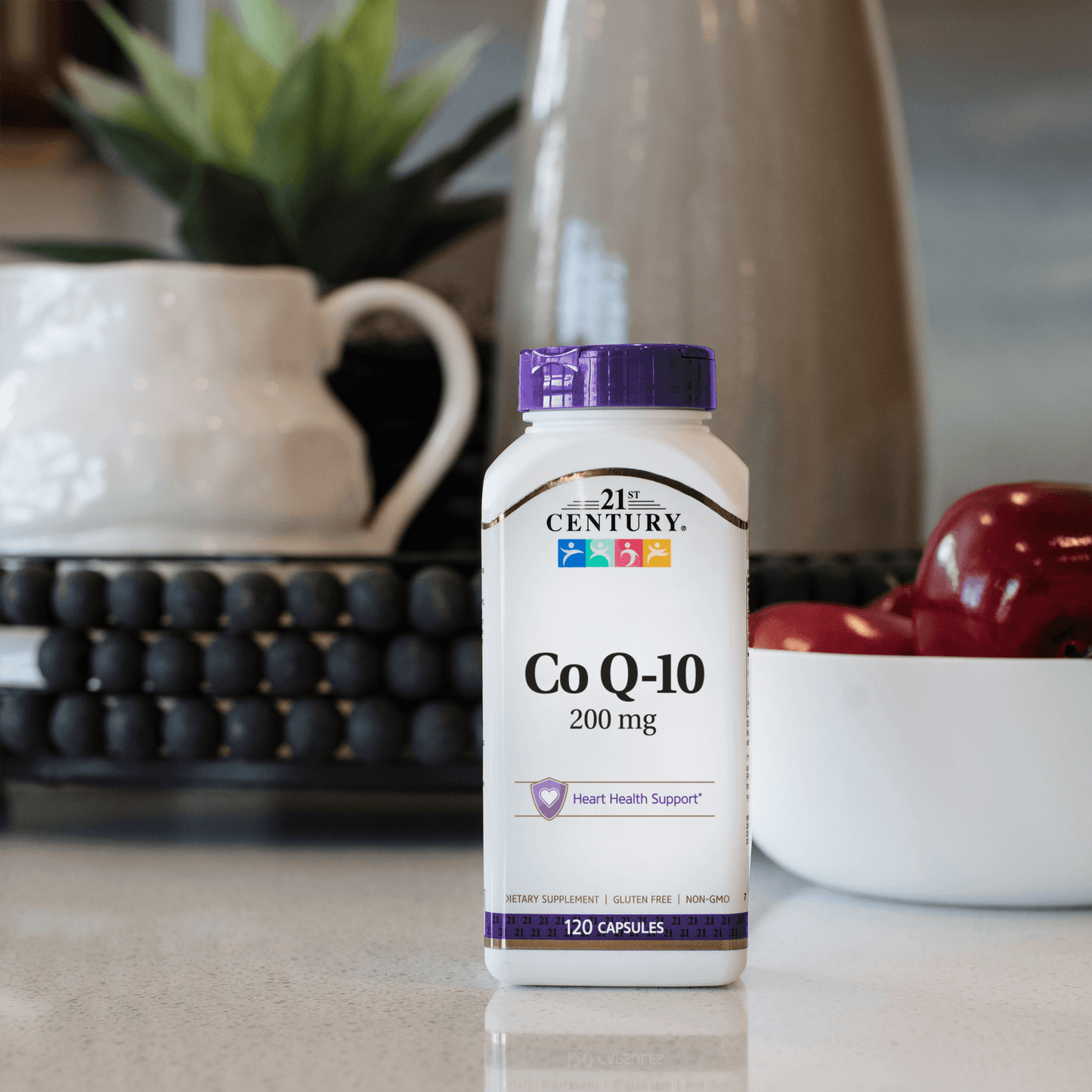A bottle of 21st Century Co Q-10 200 mg capsules sitting on a kitchen counter with a bowl of apples, a potted plant and vase in the background.