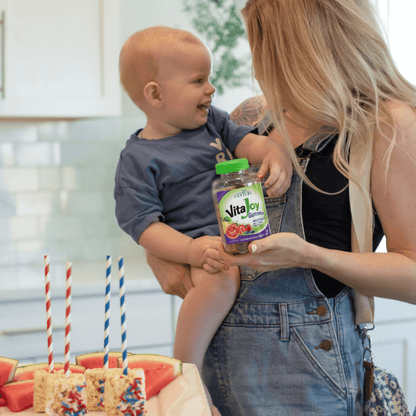 A mom holding her toddler boy with a bottle of 21st Century VitaJoy Adult Multi gummies in her hand in the kitchen.