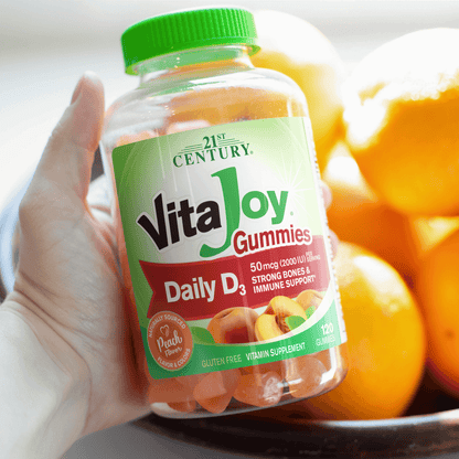 A woman holding a bottle of A bottle of 21st Century VitaJoy Daily D3 50 mcg gummies and peaches in the background.
