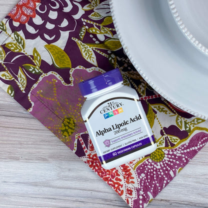 A bottle of 21st Century Alpha Lipoic Acid 200 mg capsules on a dinner napkin next to a dinner plate.