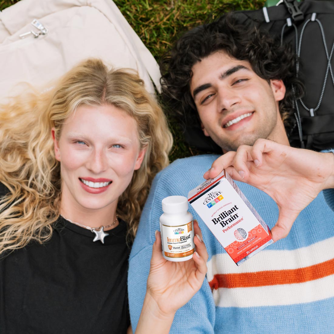 Two young adults lying on grass outdoors, smiling and holding a small white bottle and a red-and-white box of Brilliant Breathe air freshener. A backpack and clothing are visible in the background.