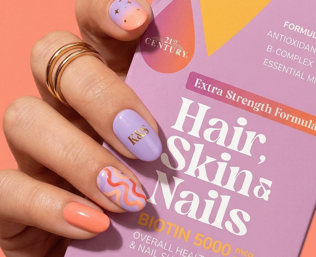 A hand with pastel lavender and orange nail art holds a box of Kalos & Splendor Biotin 5000 mcg Hair, Skin & Nails against a solid peach background.