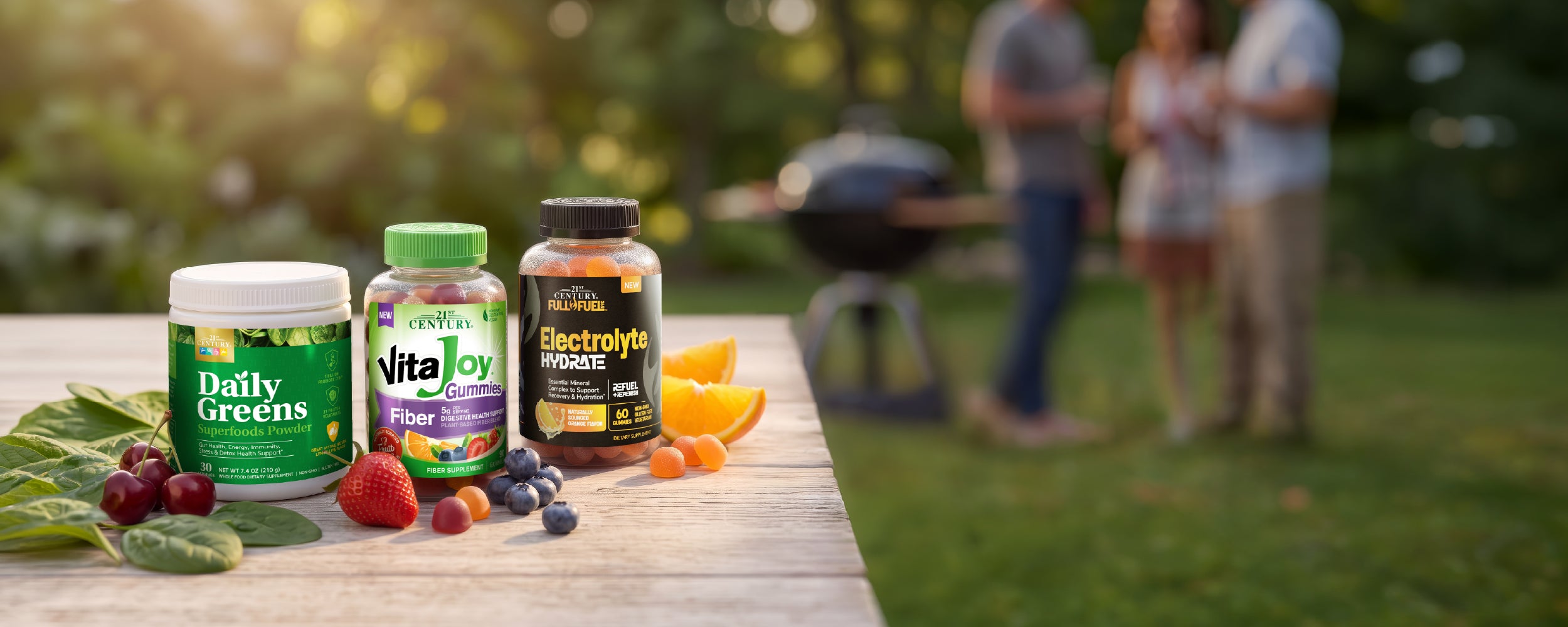 Three bottles of 21st Century supplements (Daily Greens, VitaJoy Fiber, and Electrolyte Hydrate) arranged on a wooden table outdoors with fresh fruits like strawberries, blueberries, and oranges, set against a blurred background of a backyard grill.