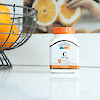 A bottle of 21st Century Vitamin C 500 mg tablets on a counter with a wire bowl of oranges to the left.