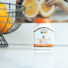 A bottle of 21st Century Vitamin C 500 mg with Rose Hips tablets sitting on a kitchen counter next to a wire bowl of oranges.