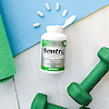 A bottle of 21st Century Sentry Senior Adults 50+ Multivitamin and Multimineral laying on a yoga mat with tablets coming out of the top and dumbbells below.