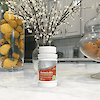 A bottle of 21st Century One Daily Women's 50+ Multivitamin and Multimineral tablets sitting on a kitchen counter with a vase of flowers and jars of fruit in the background.