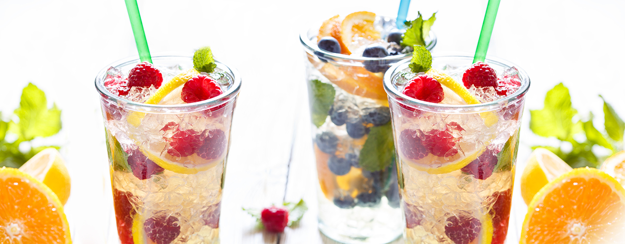 Two glasses with straws filled with crushed ice, lemons, limes, blueberries and raspberries.