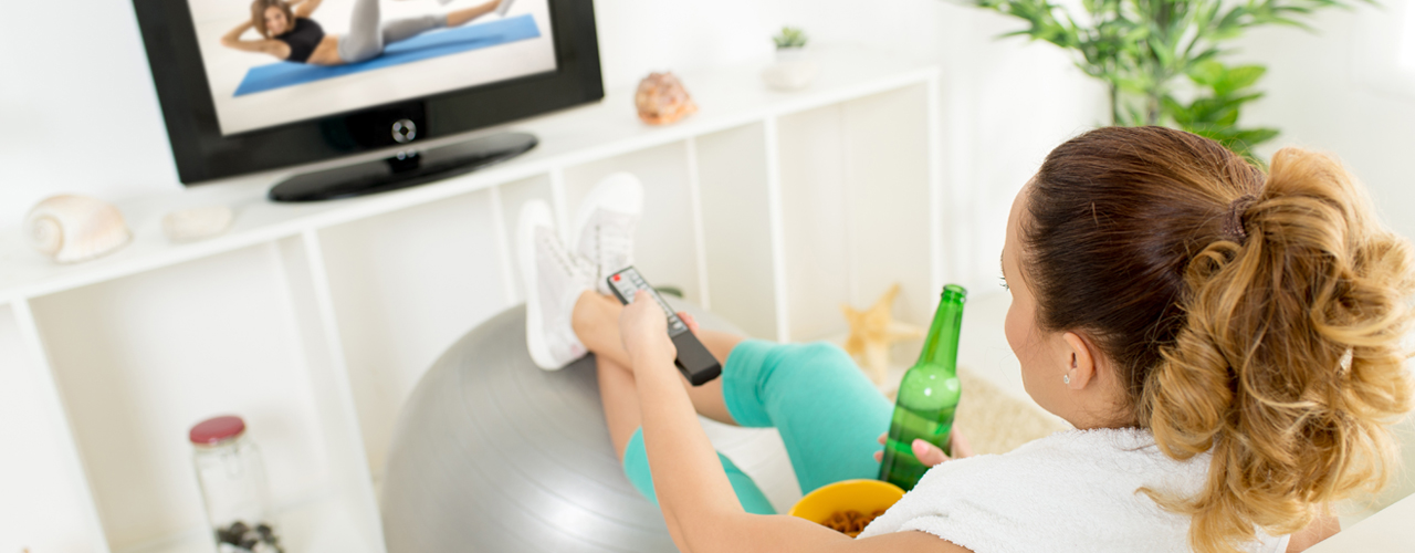 A girl in workout wear sitting on the couch drinking soda, eating snacks and watching TV.