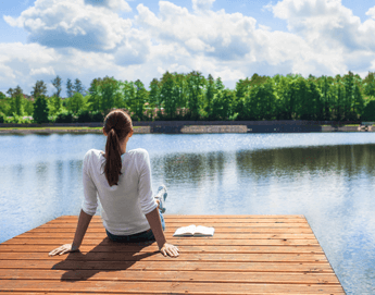 A back view of a girl sitting on a dock looking out at the calm water.