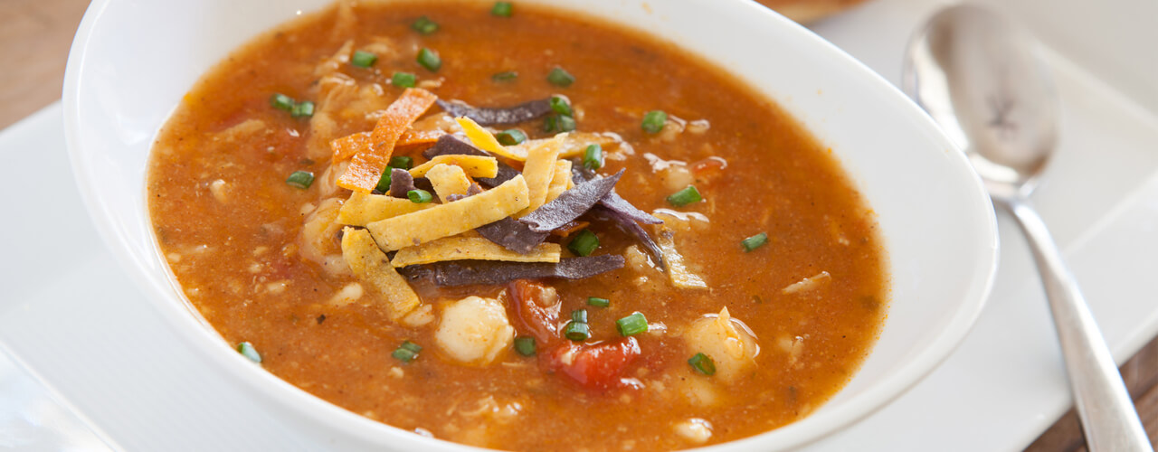 A bowl tortilla soup with spoon and bread roll.