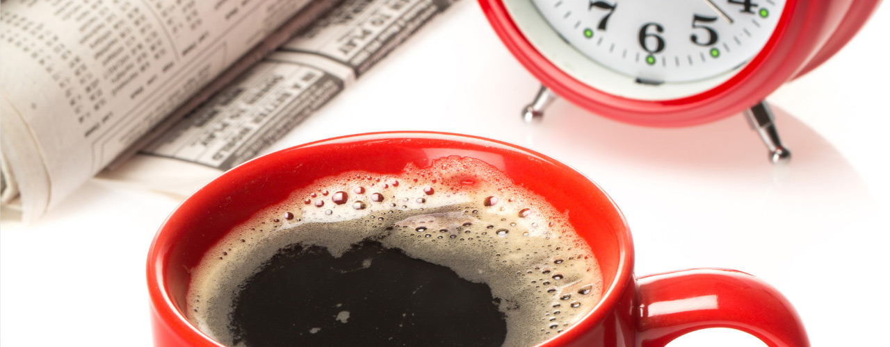 A red coffee cup filled with coffee next to an analog clock and newspaper.