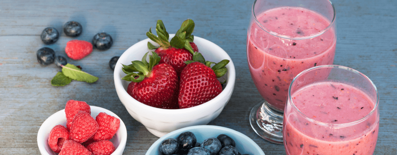 Two glasses filled with smoothies next to bowls of strawberries, blueberries and raspberries.